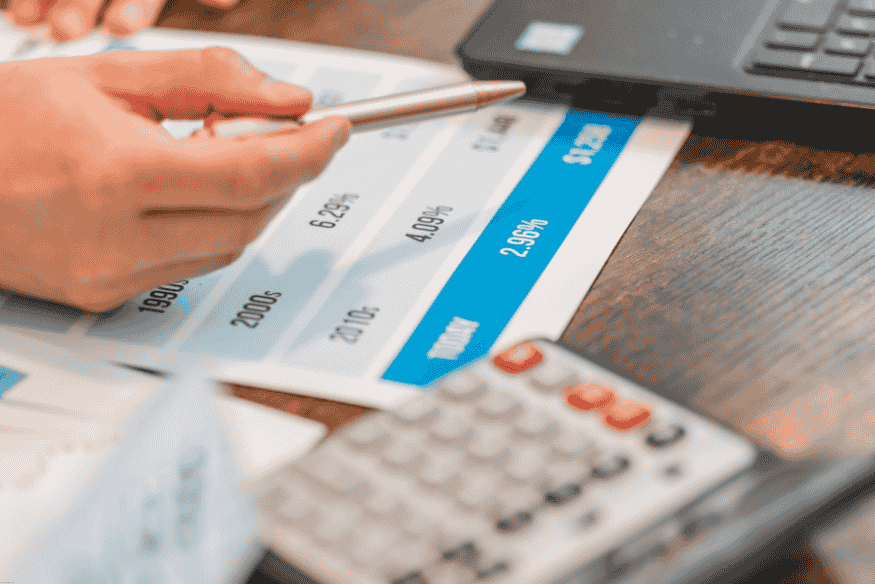 Hand holding a pen over printed interest rate charts next to a calculator and laptop.