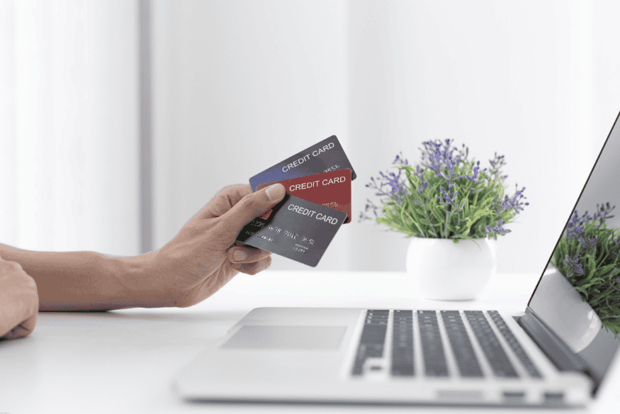 Hand holding multiple credit cards in front of a laptop on a white desk